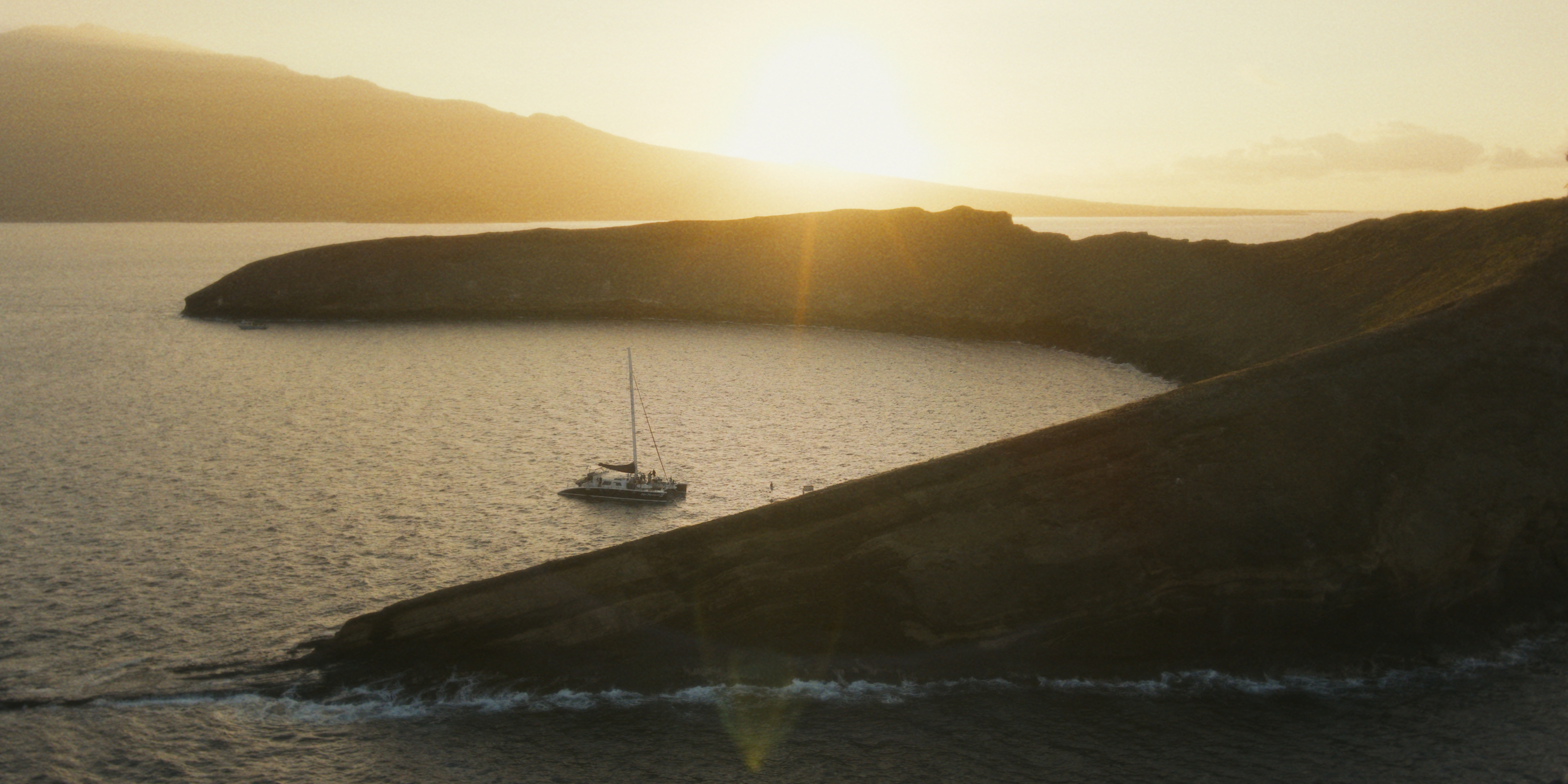 A catamaran crossing sunlit water beneath island ridgelines.